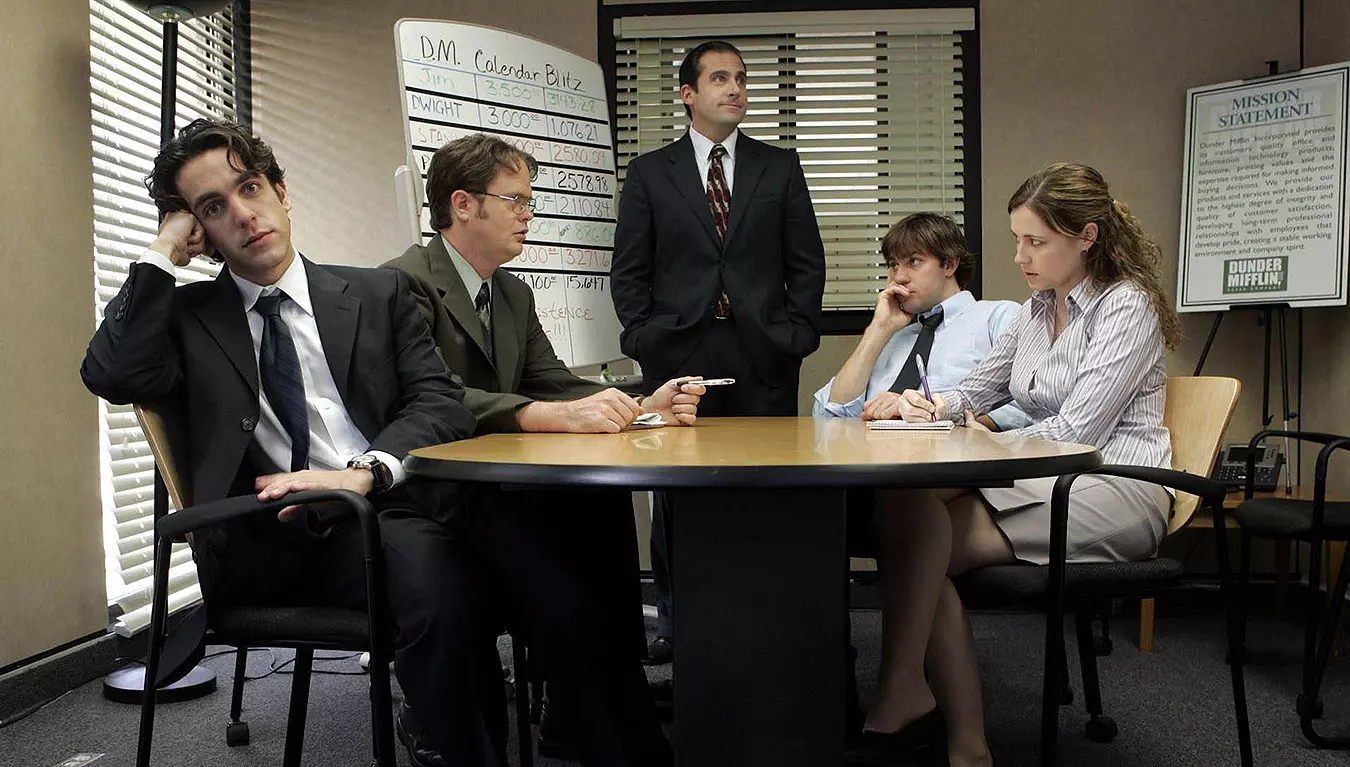 Escena de serie de comedia en oficina con empleados reunidos alrededor de una mesa en ambiente corporativo