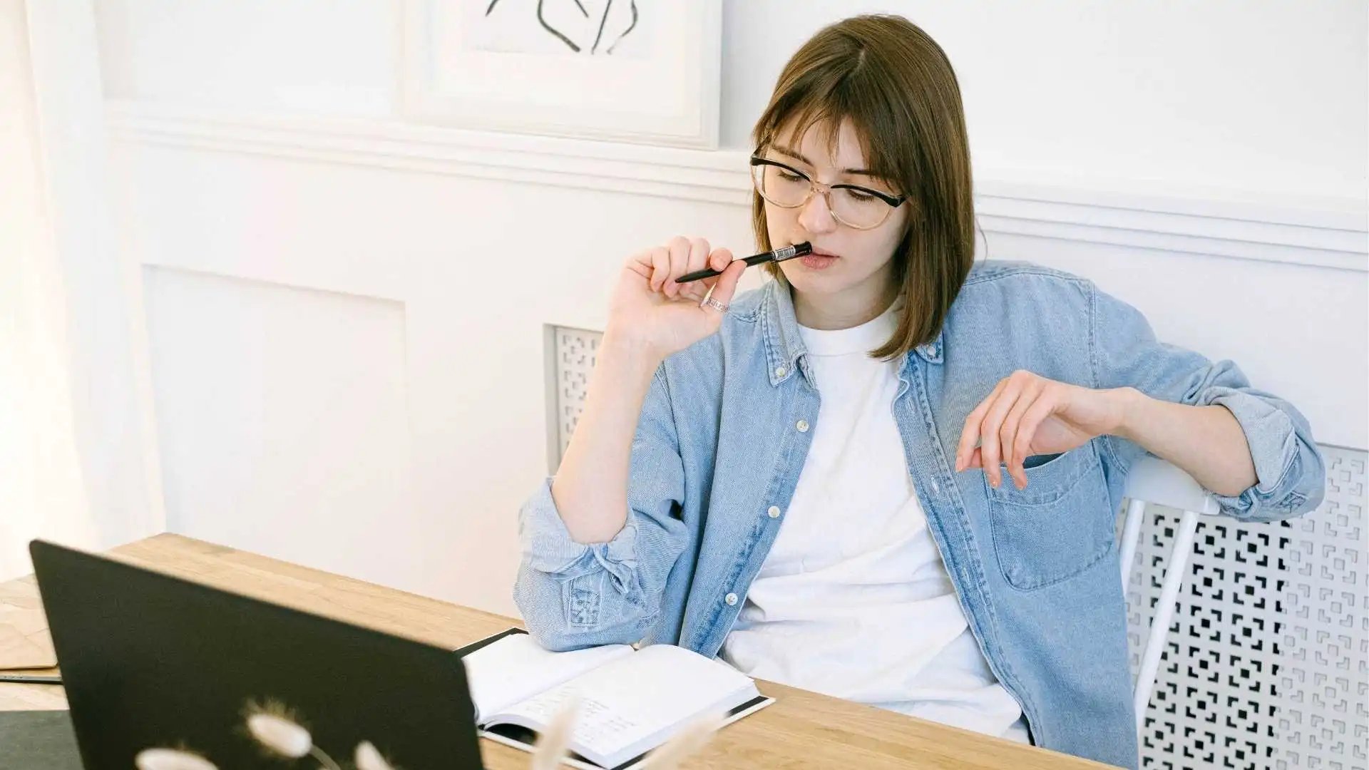 Mujer joven pensando frente a una laptop mientras escribe ideas en una libreta, representando el proceso creativo del naming y la creación de un nombre de marca.