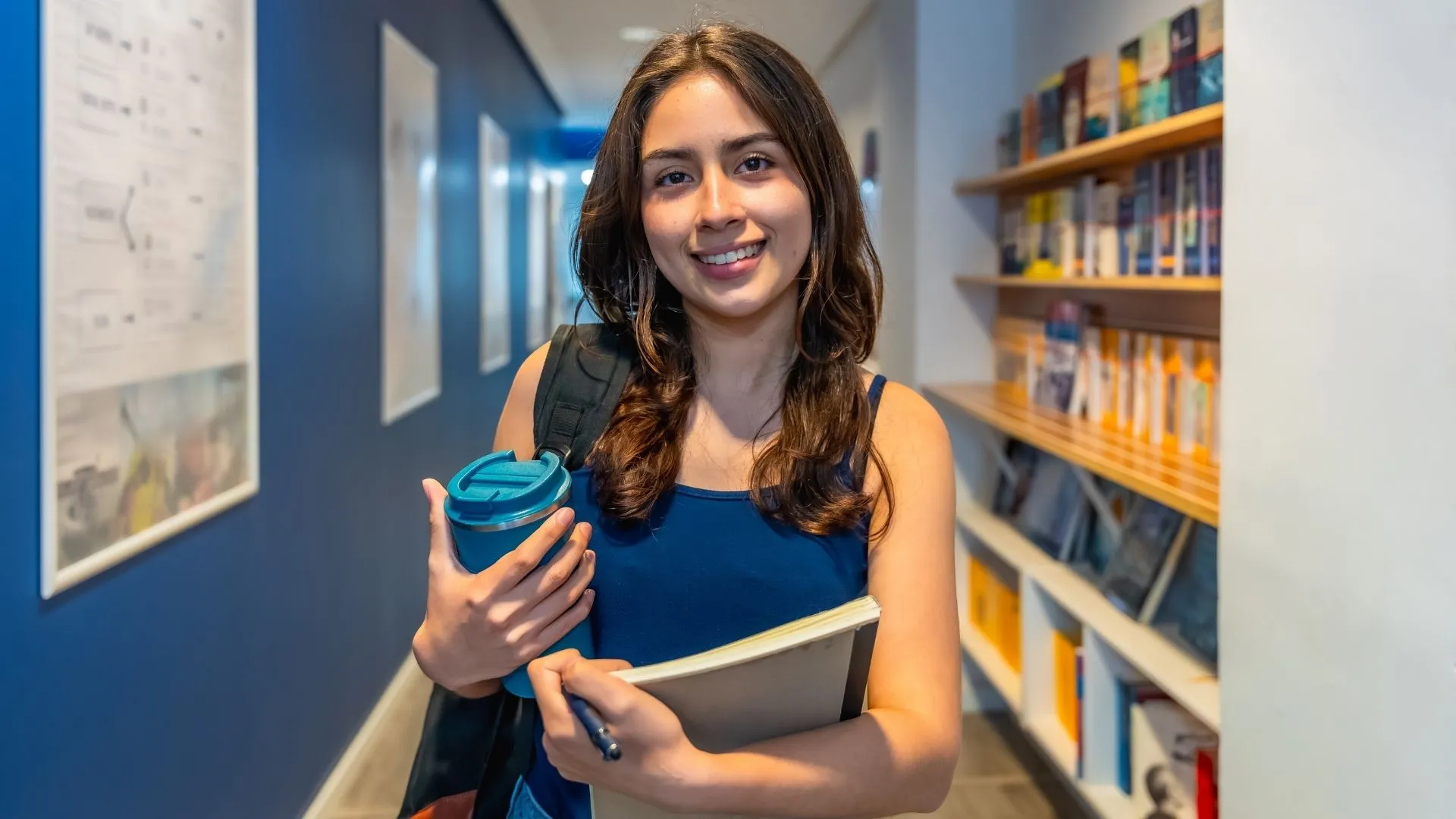 Mujer estudiante joven, de temperamento alegre, sosteniendo un termo de café, así como un libro, en un pasillo de la universidad.