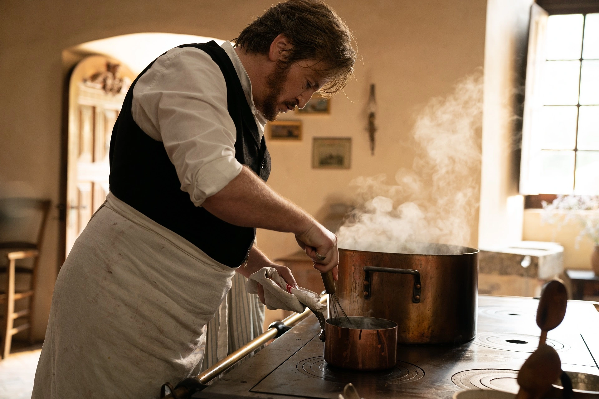 Chef removiendo una preparación en una cacerola de cobre junto a una olla humeante.
